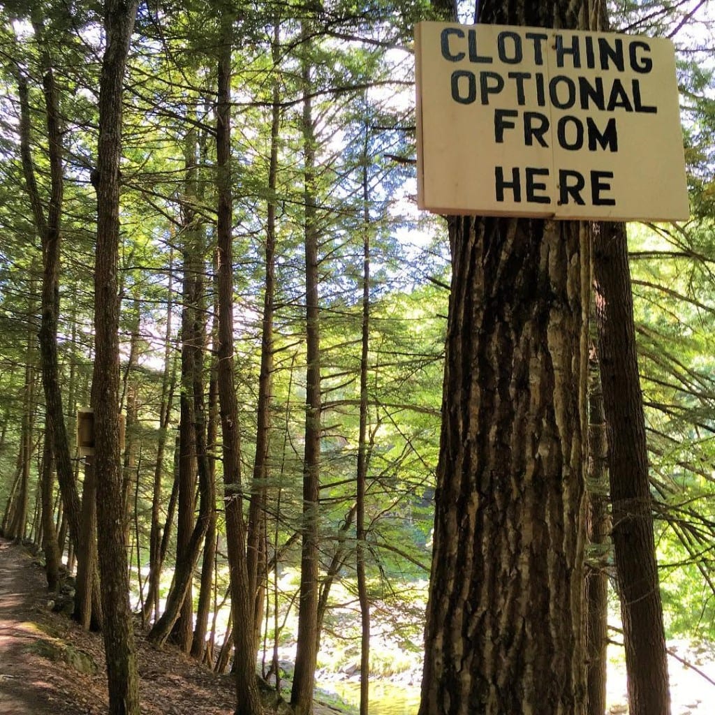 Cream-colored trail sign on a tree in a sunlit Vermont forest reading Clothing Optional From Here — path and glimpse of rocky river at Rock River, Newfane