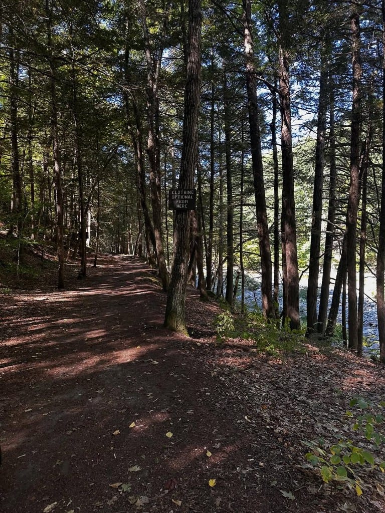 Shaded dirt trail at Rock River, Vermont, with a tree-mounted sign reading Clothing Optional From Here — rocky river visible through the trees