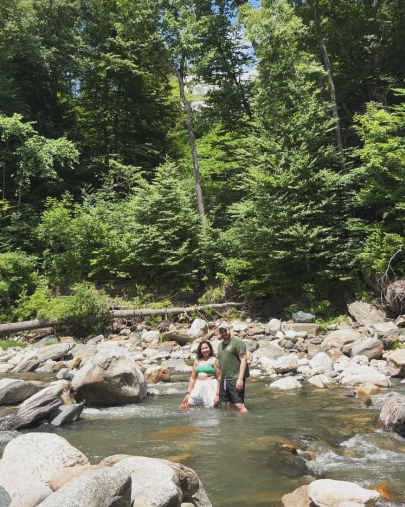 A couple smiles while wading in clear shallow Rock River water in Newfane, Vermont — smooth stones, log on the bank, lush green trees