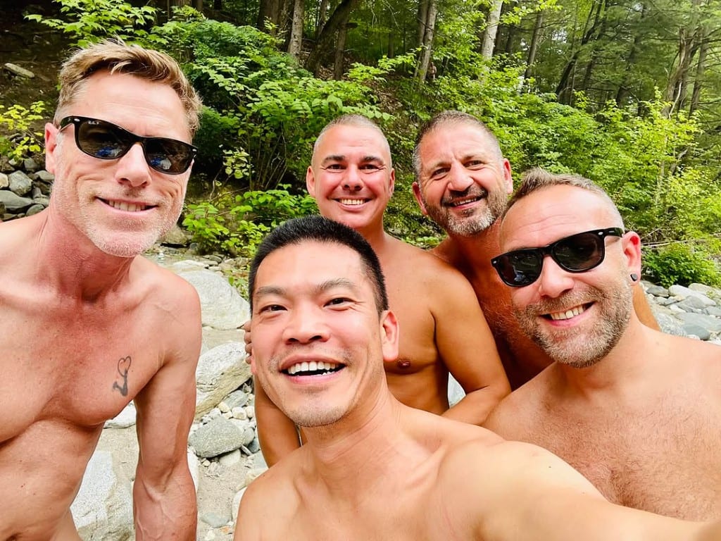 Five visitors smiling together on sun-warmed river stones at Rock River, Vermont — shirts off, summer forest behind, friendly group moment in Windham County