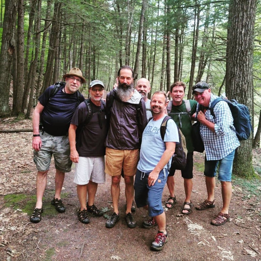 A group of seven hikers smiling together on a wooded trail near Rock River, Vermont — day packs, summer forest, friendly trail day