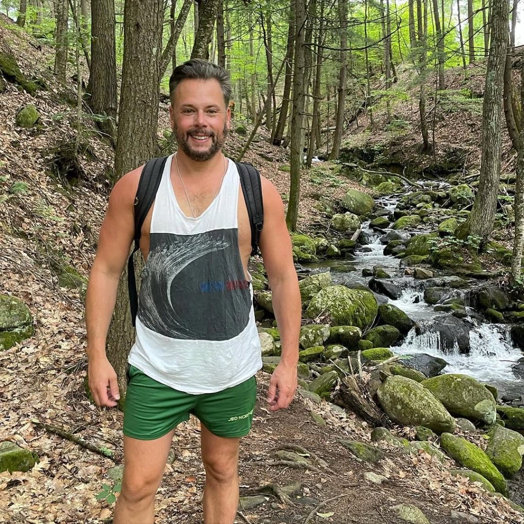 Smiling hiker with a pack beside a small clear stream on a leafy trail at Rock River, Vermont — green woods, summer day