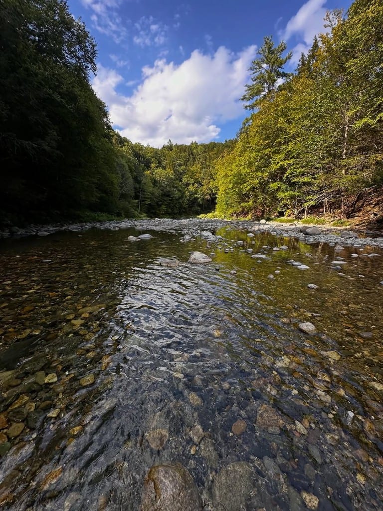 Clear shallow Rock River flowing over stones toward a forested ridge, Vermont — bright blue sky with clouds, sunlit and shaded banks