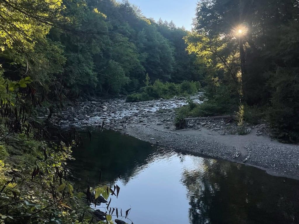 Golden-hour sunburst over a calm bend of Rock River, Vermont — dark pool reflecting sky, rocky shallows, sandy beach and dense green forest