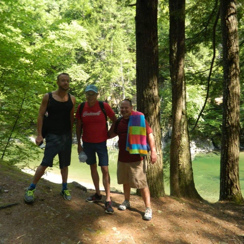 Three friends on a wooded trail above emerald-green Rock River water in Vermont — towels and packs, summer outing
