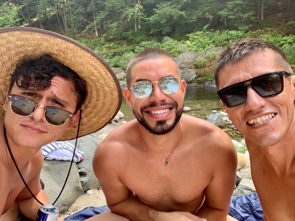 Three visitors smiling together on sun-warmed river rocks beside clear water at Rock River, Newfane, Vermont — sunglasses, lush green forest across the bank