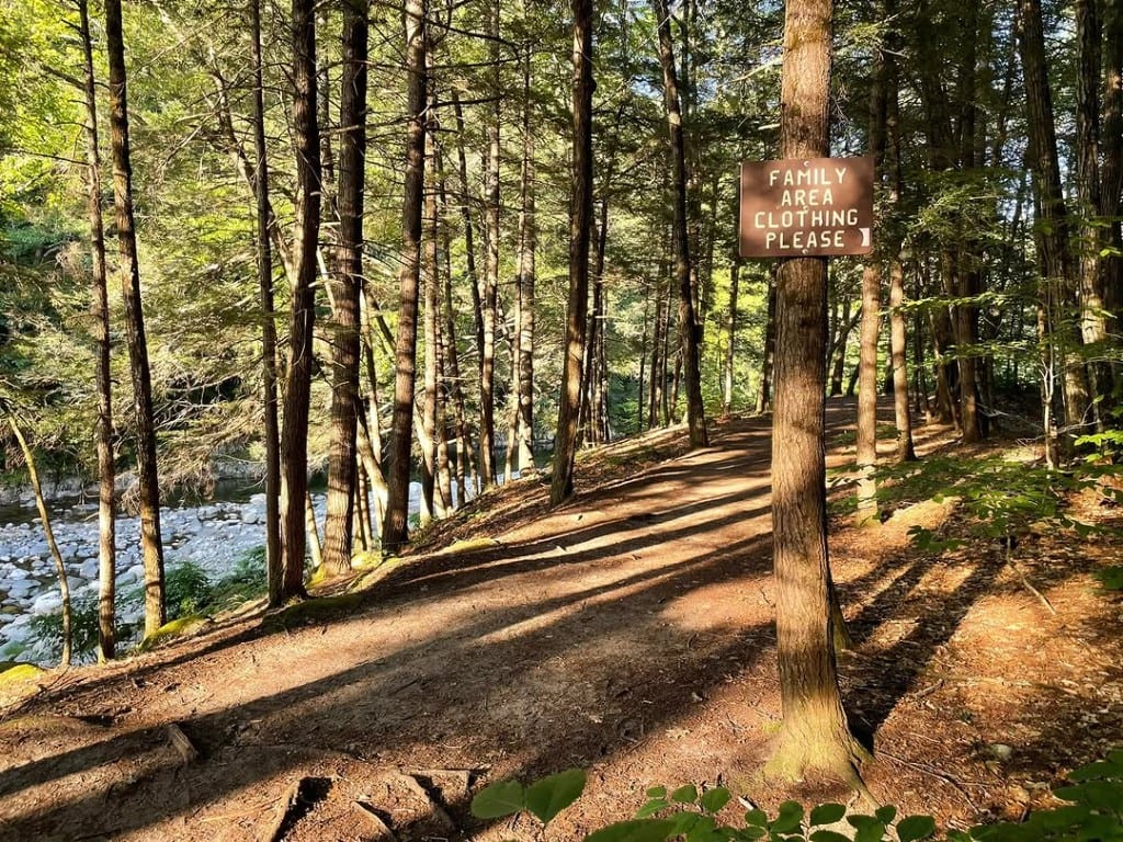 Sunlit forest trail beside the Rock River in Vermont with a brown sign on a tree reading Family Area Clothing Please and an arrow along the path