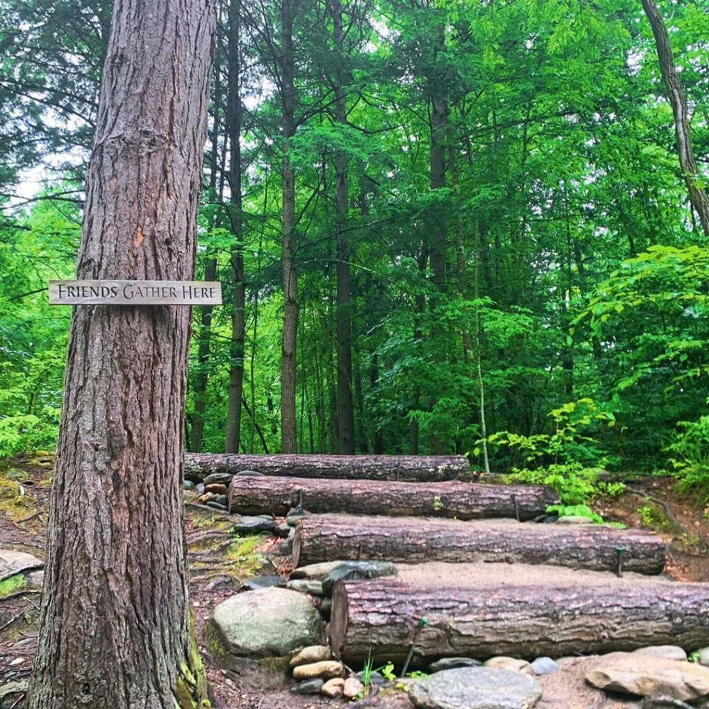Wooden stairs of sand and stone leading into the woods at Rock River, Vermont — hand-painted sign on a tree reads Friends Gather Here