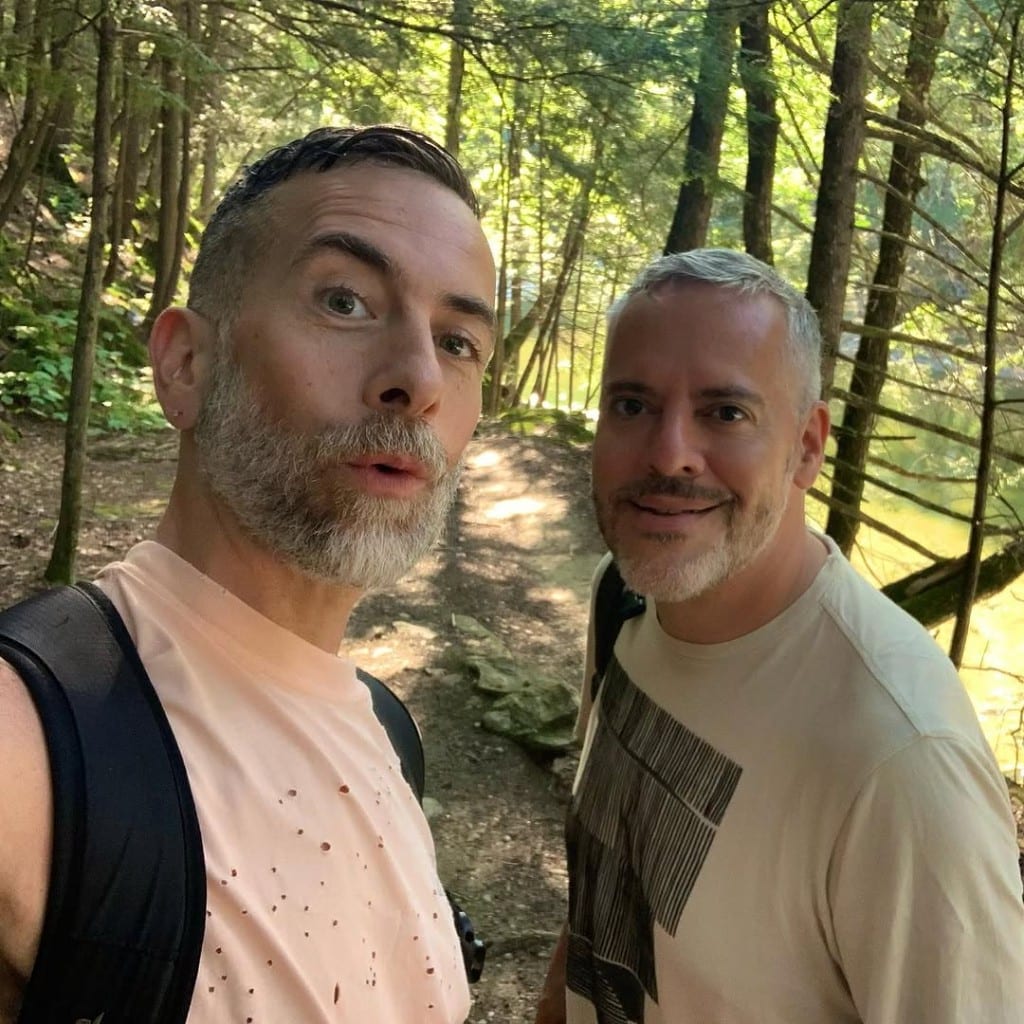 Two friends on a sun-dappled forest trail at Rock River, Vermont — hiking packs, warm smiles, glimpse of bright river through the trees