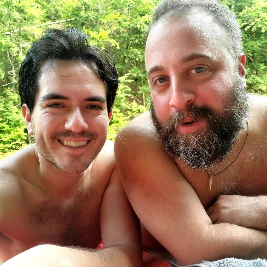 Two visitors relaxing shirtless on towels at the riverbank, Rock River, Vermont — warm smiles, soft-focus summer forest behind