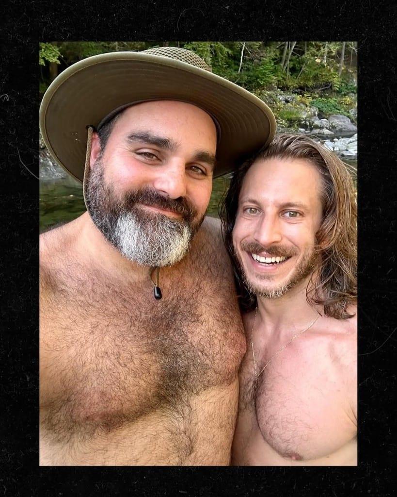 Two smiling visitors pose for a selfie beside a shallow rocky Rock River in Newfane, Vermont — sun hats, clear water, summer trees