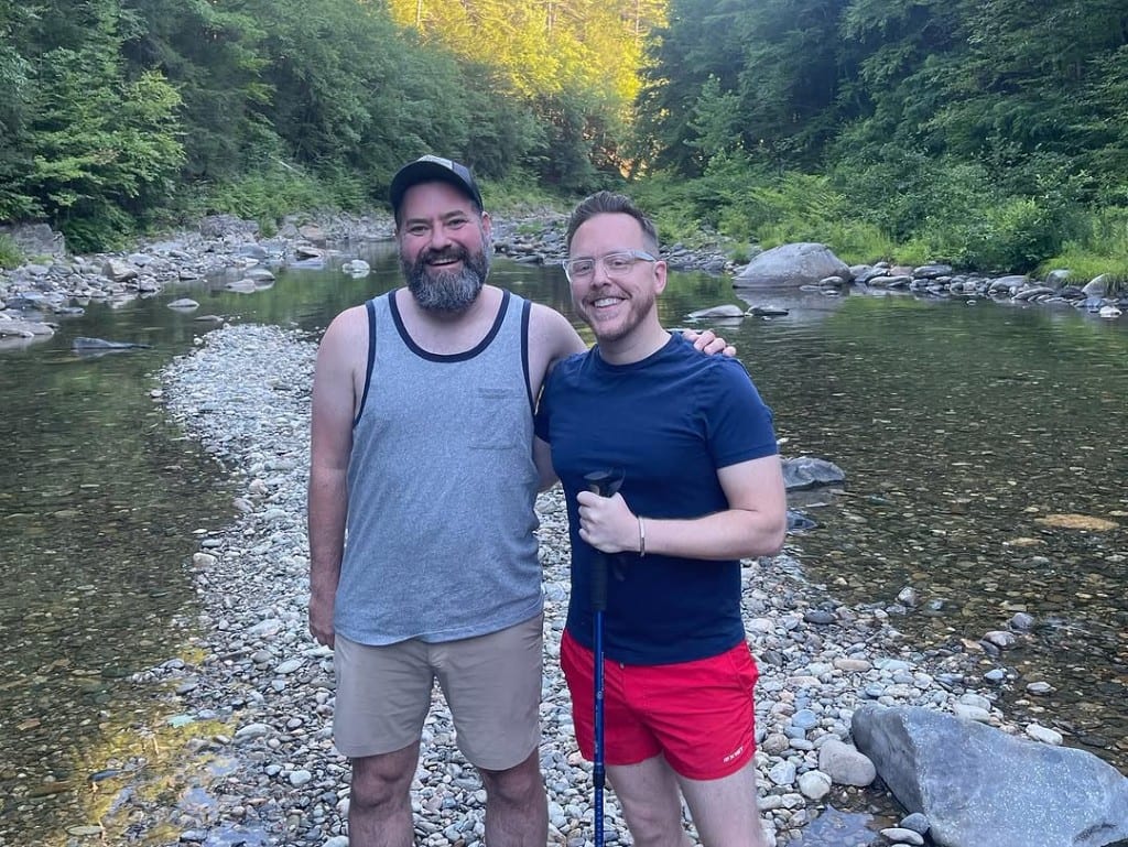 Two visitors smiling on a rocky bar in the shallow Rock River, Newfane, Vermont — clear water, lush green forest, relaxed summer outing