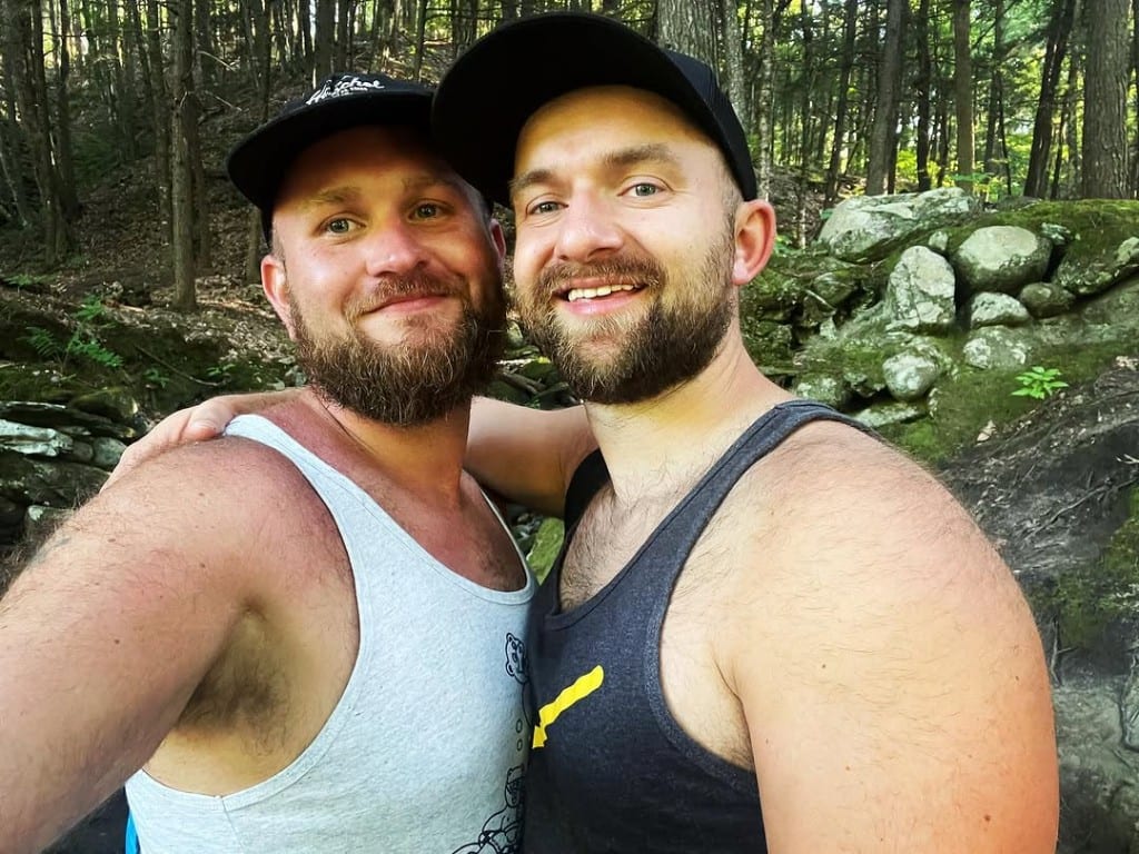 Two visitors in caps and tank tops smiling together in the woods at Rock River, Vermont — mossy boulders and dense green trees behind