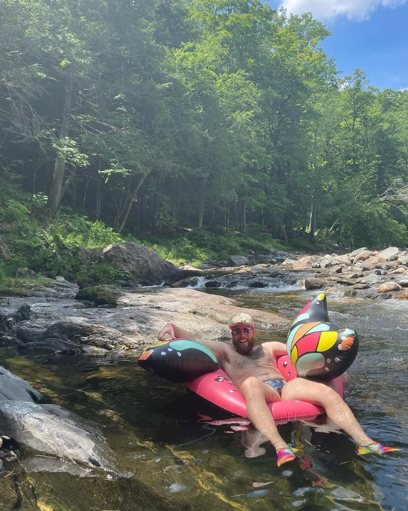 A smiling visitor floats in a bright pink tube on clear shallow Rock River water in Vermont — forested banks and summer sky