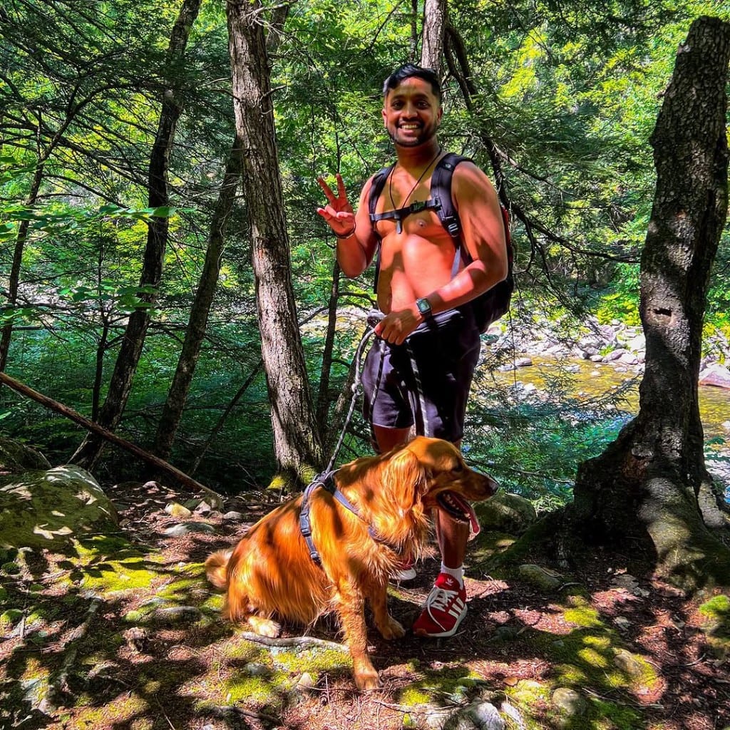 A smiling visitor with a golden retriever on a leash on a sunlit forest trail near Rock River, Vermont — rocky river glinting through the trees