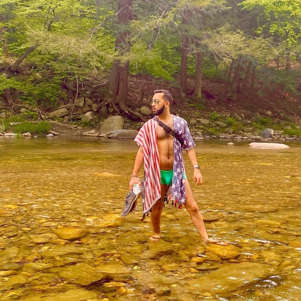 Visitor standing in shallow clear water at Rock River, Vermont — bright swimwear, open shirt, sneakers in hand, rocky bottom and green forest