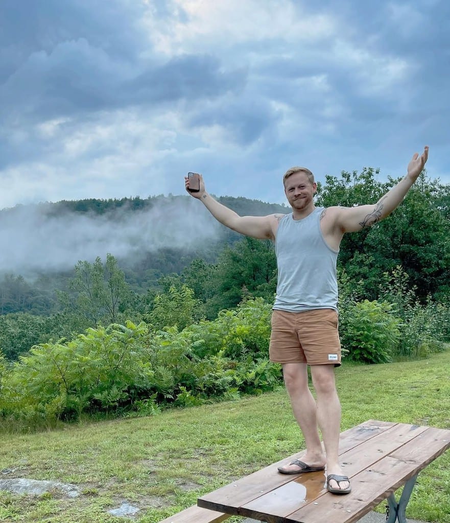 Visitor with arms wide on a wet wooden picnic table, misty forested hills behind — celebratory outdoor moment, Rock River area, southern Vermont