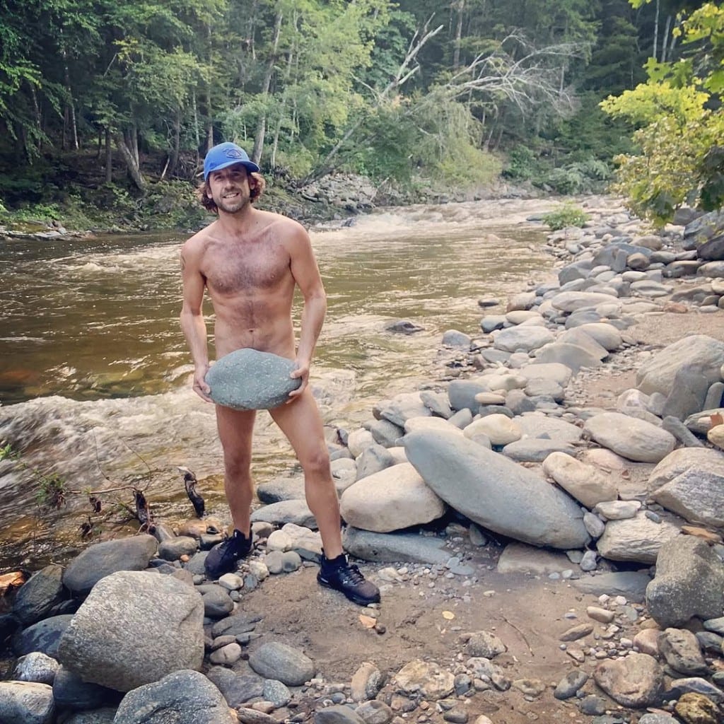 Playful visitor on the rocky Rock River bank, Vermont, smiling and holding a smooth flat stone in front — forested river scene, lighthearted river day