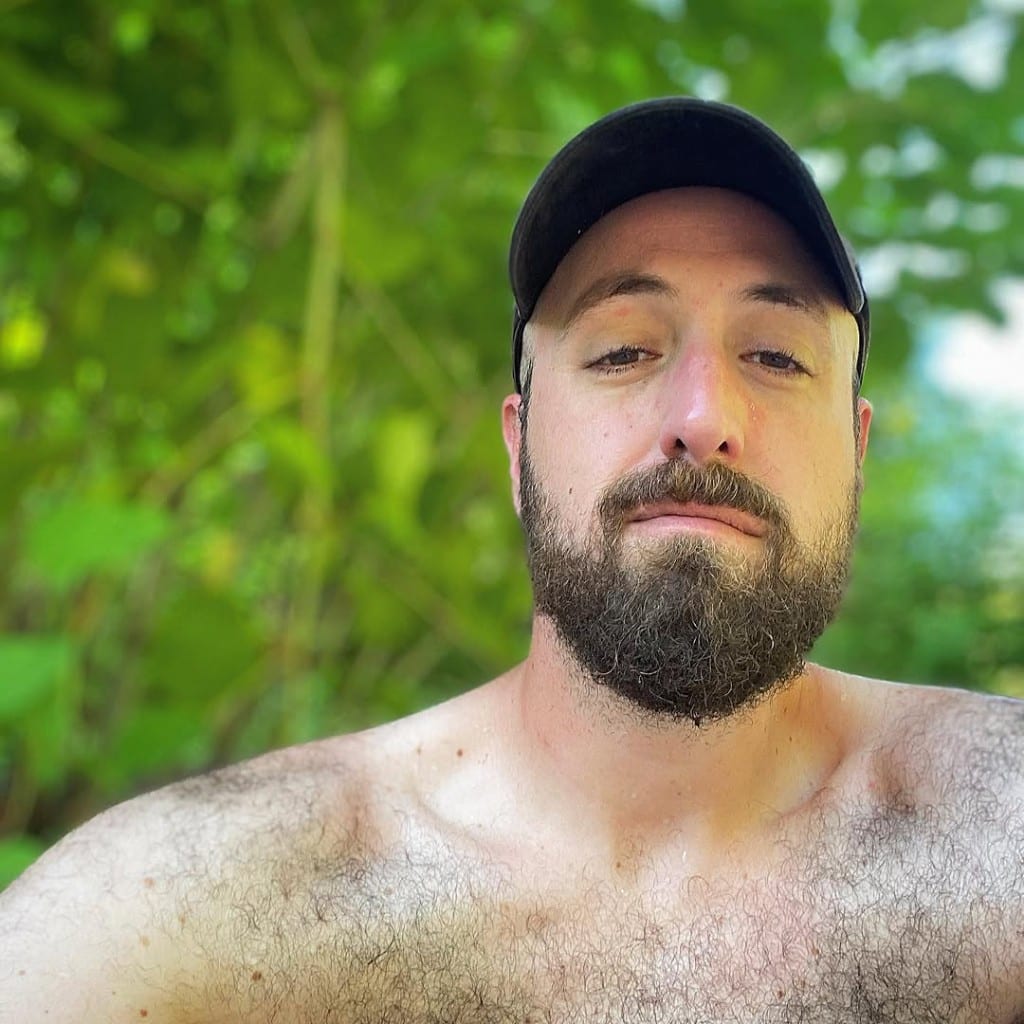 Close-up portrait of a visitor in a black cap, shirtless after a swim, with soft green forest bokeh at Rock River, Vermont