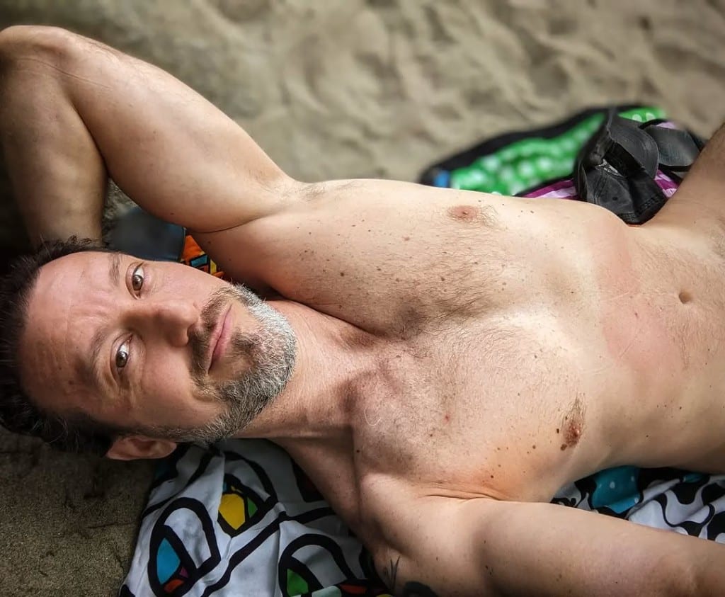 Visitor relaxing on a patterned towel on sandy riverbank at Rock River, Vermont — calm smile, bright summer day