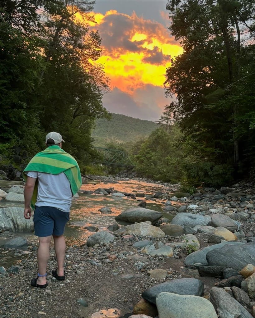 A visitor with a green striped towel on their shoulders stands on the rocky bank of Rock River, Vermont, at sunset — green bridge and fiery orange sky