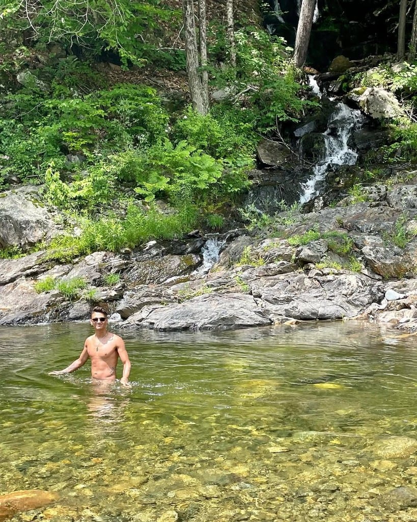 A visitor stands in a clear green swimming hole at Rock River, Vermont, with a small waterfall and ferns on the rocky bank behind