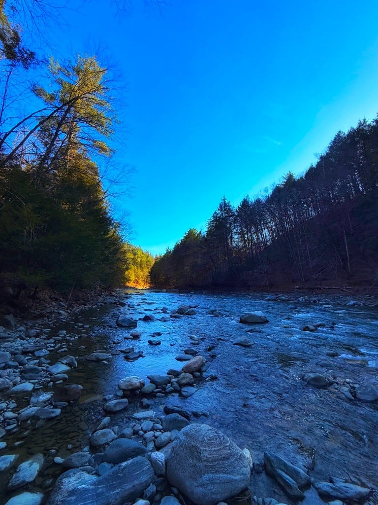 Rock River at dusk — Newfane, Vermont Evening light over the Rock River in Newfane, Vermont — rounded cobbles, shallow current, and dark conifer ridges under a deep blue sky