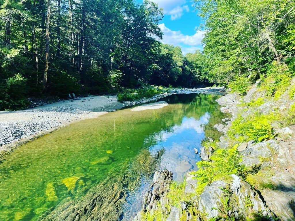 Rock River Vermont swimming hole hero — Newfane VT Serene summer view of Rock River in Newfane, Vermont — clear green-tinted water, a sandy beach, rocky shore, and forest under a blue sky