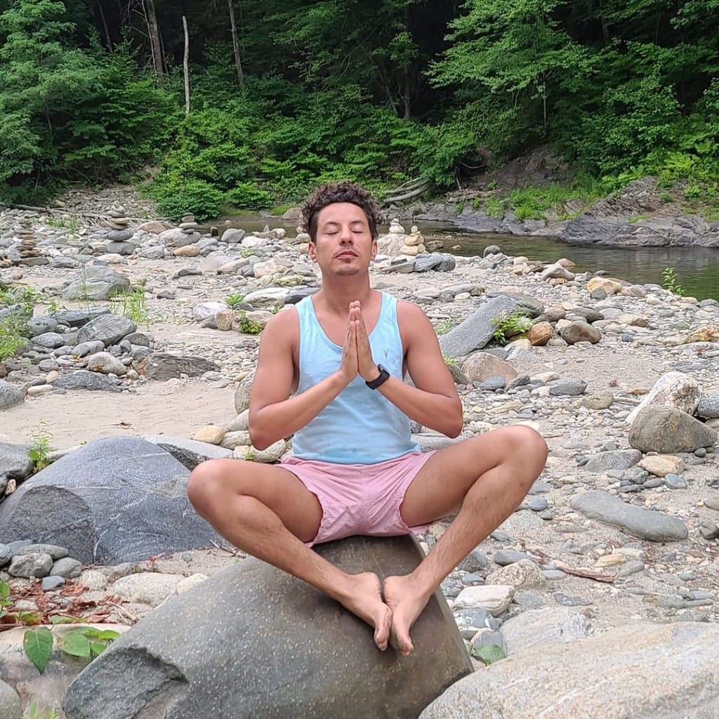 Rock River — quiet moment on the river Visitor meditating on a large boulder in a rocky Rock River bed, Vermont — hands at heart, small stone cairns and green forest in the background